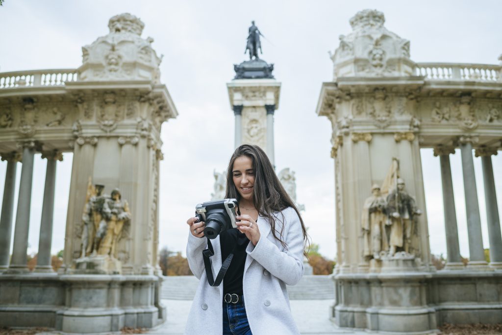 Una mujer con una sonrisa en el rostro se encuentra de pie frente al monumento de Alfonso XII en el hermoso Parque del Retiro de Madrid, sosteniendo una cámara.